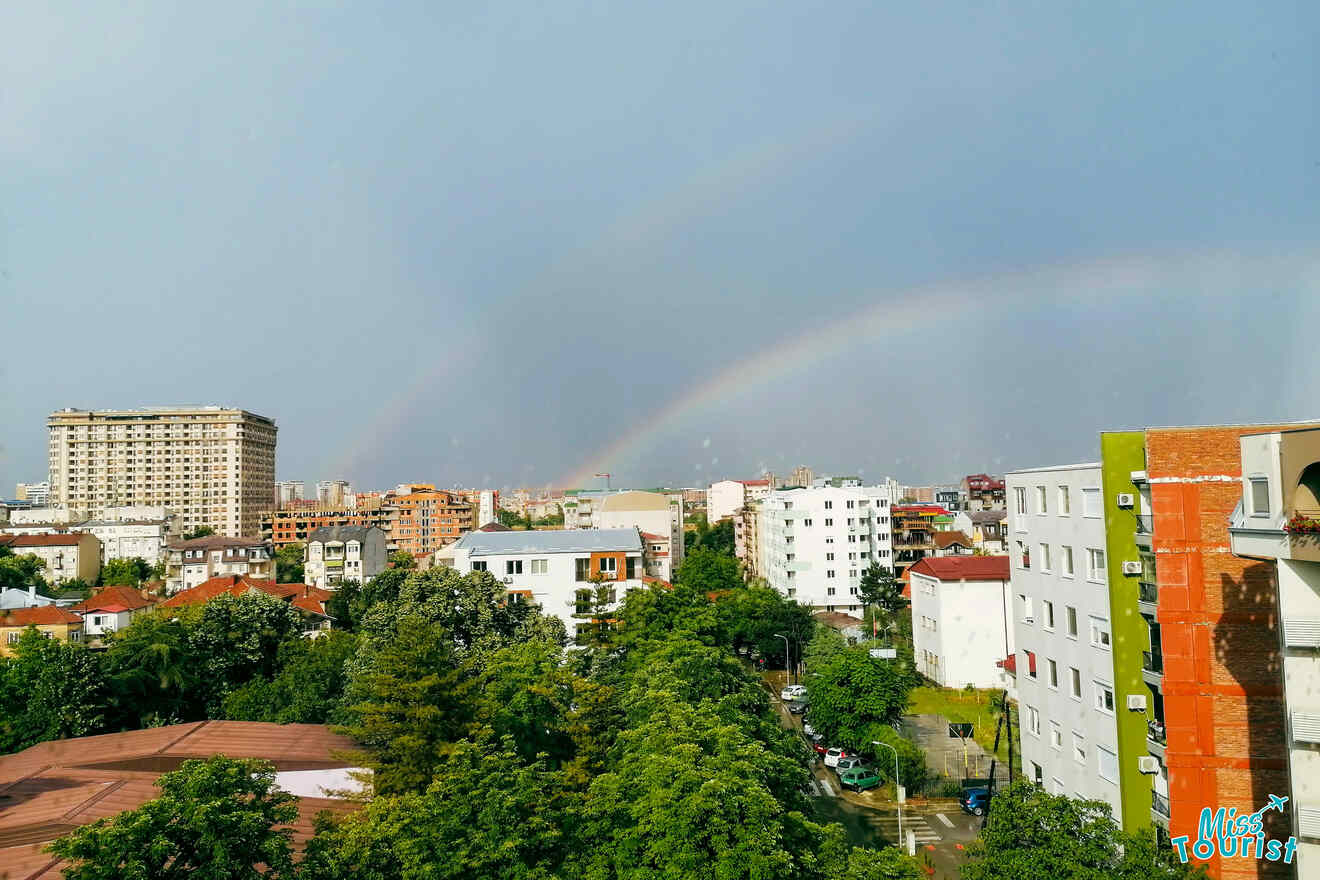 A cityscape featuring a mix of tall and mid-rise buildings with lush green trees. A faint rainbow is visible in the sky above the skyline.