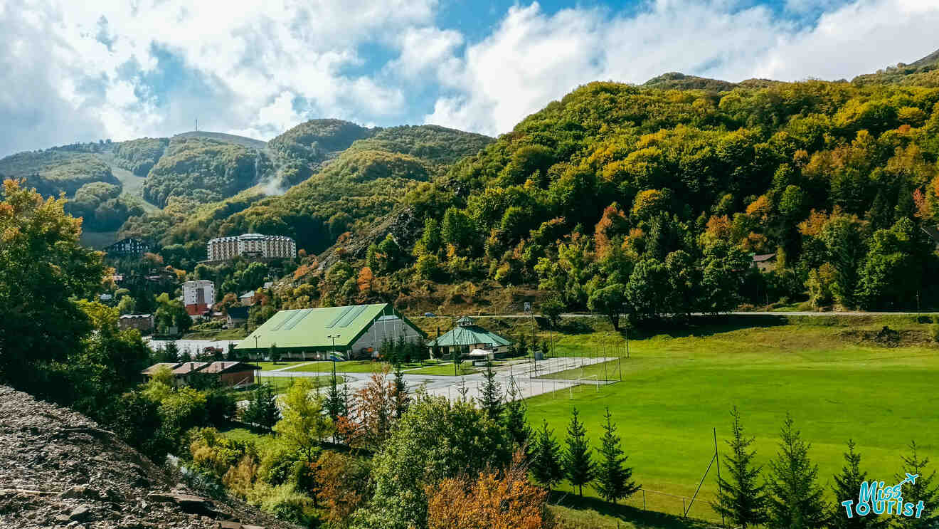 A scenic mountain valley features buildings, green fields, and tennis courts surrounded by lush, forested hills under a partly cloudy sky.