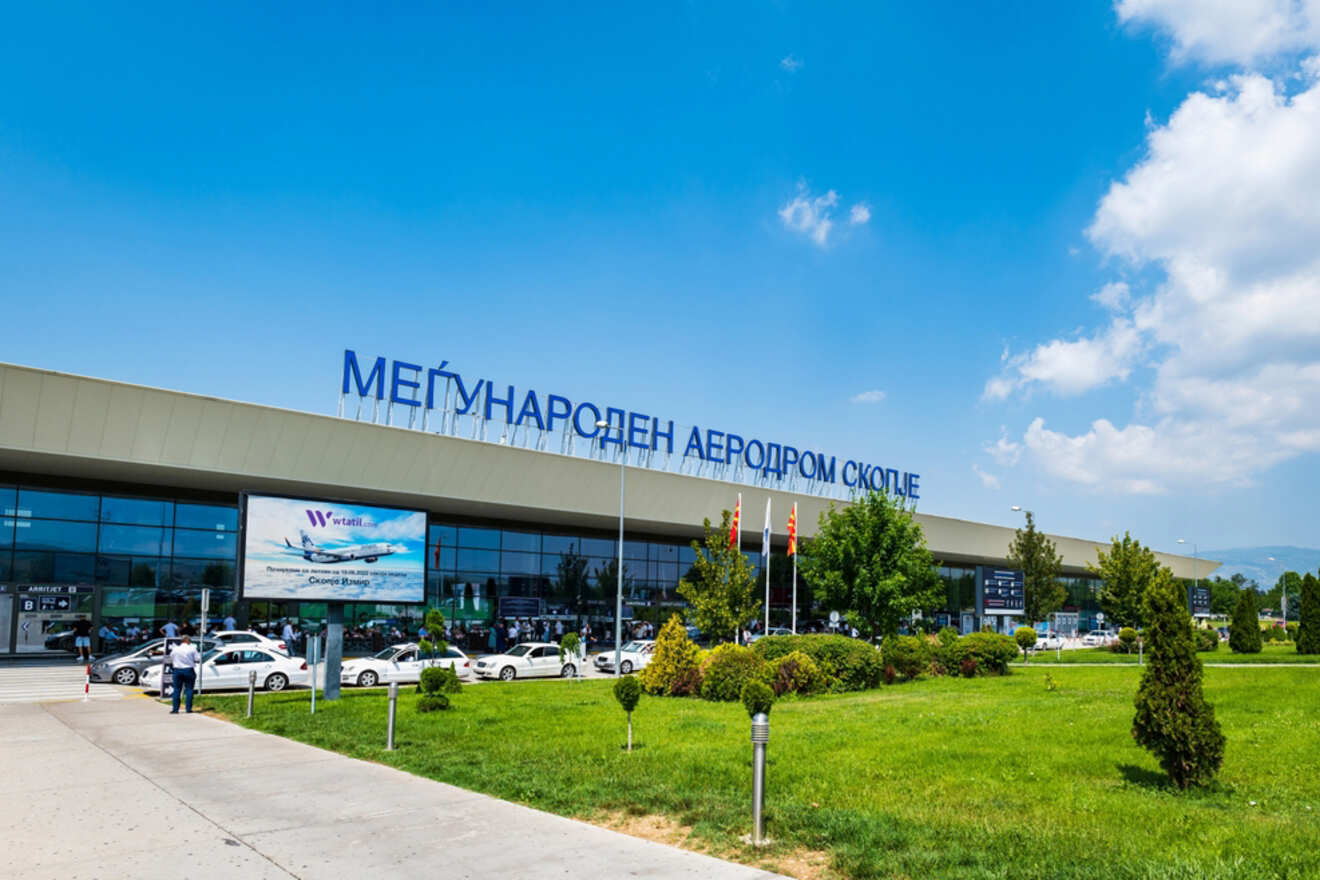 Exterior view of Skopje International Airport with visible signage, parked cars, and landscaping under a partly cloudy sky.