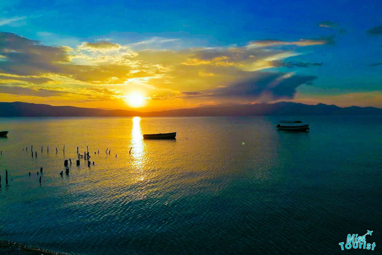 Sunset over a calm lake with two boats floating and wooden posts emerging from the water. A mix of blue, orange, and yellow hues fills the sky. 