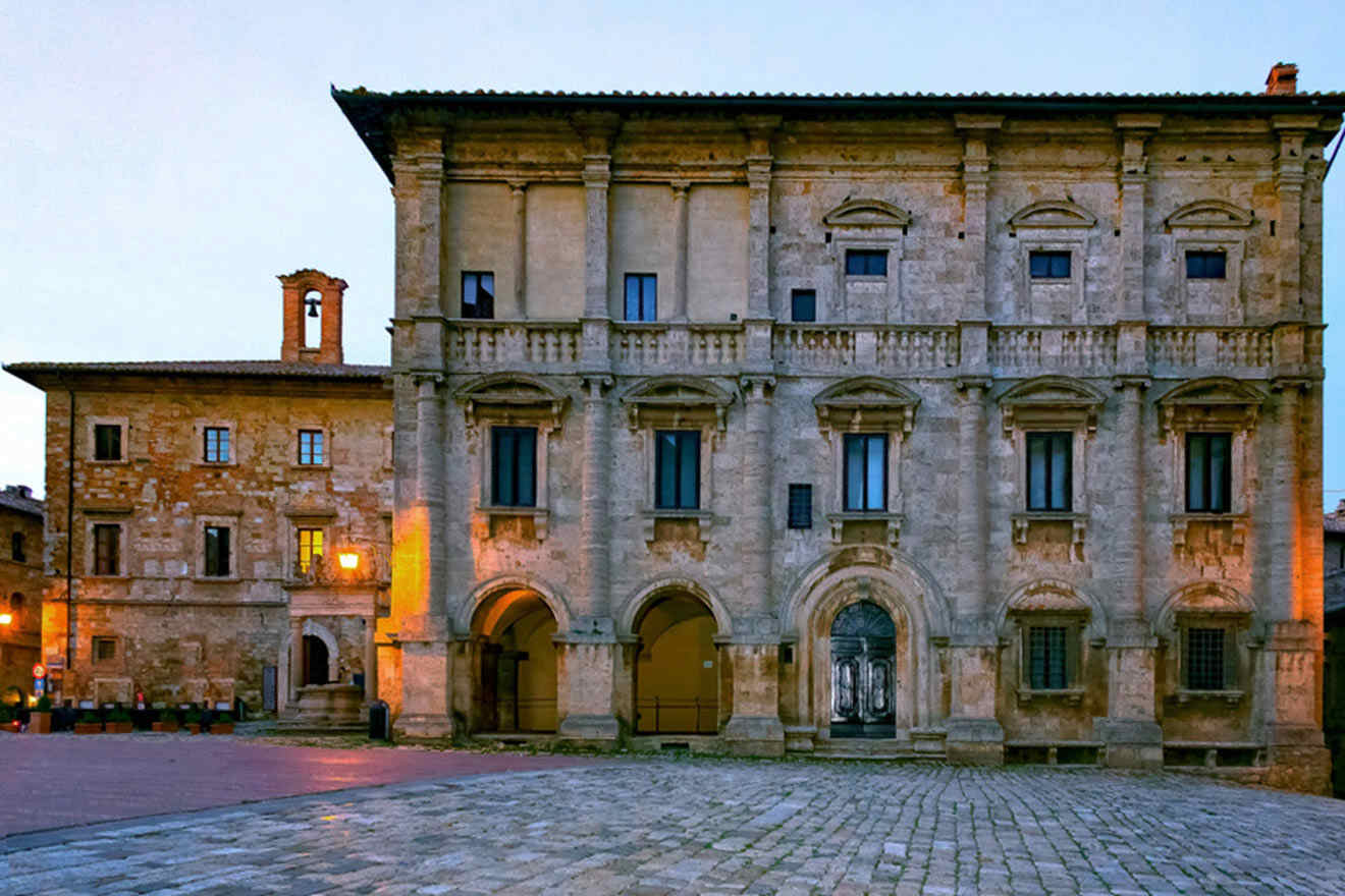 Historic stone building with arched doorways and windows, lit by streetlights at dusk. The building stands in a paved square next to another stone structure.