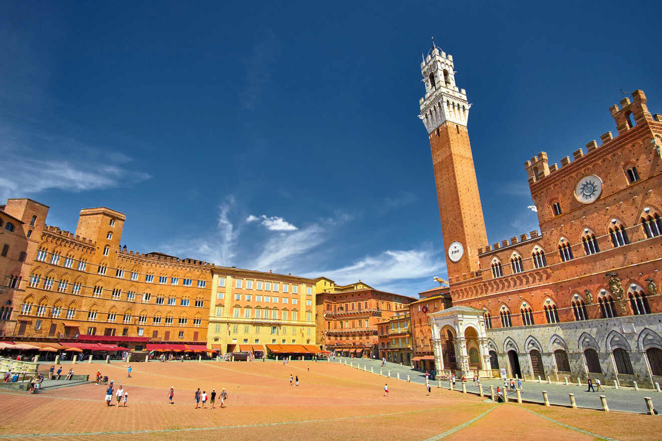 An open square in Siena, Italy, with historic buildings, including the Torre del Mangia and the Palazzo Pubblico, under a clear blue sky. People are walking and gathering in the square.