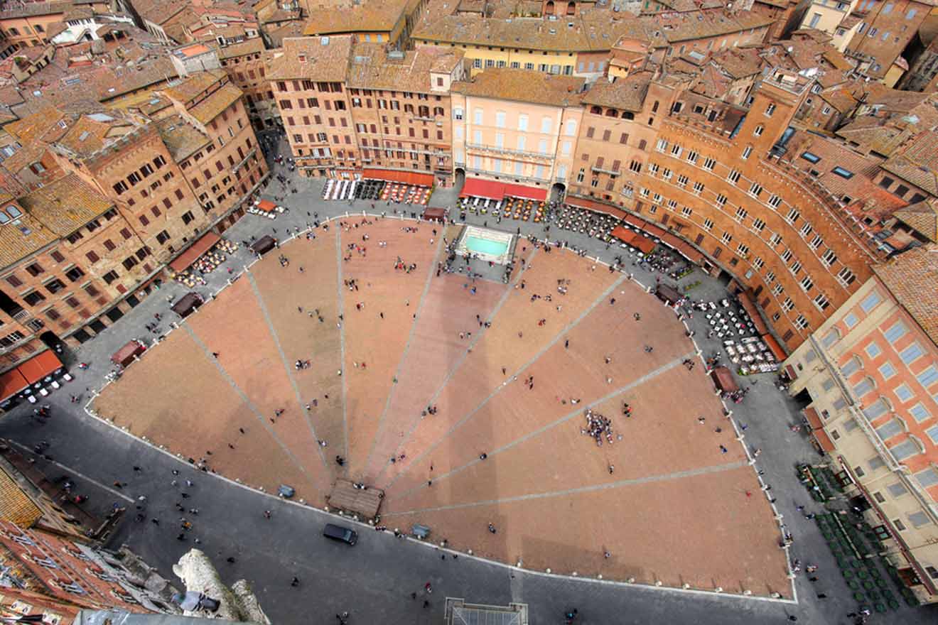 An aerial view of the Piazza del Campo in Siena, Italy, showcasing its distinctive shell shape and surrounded by historic buildings with a few groups of people scattered across the plaza.