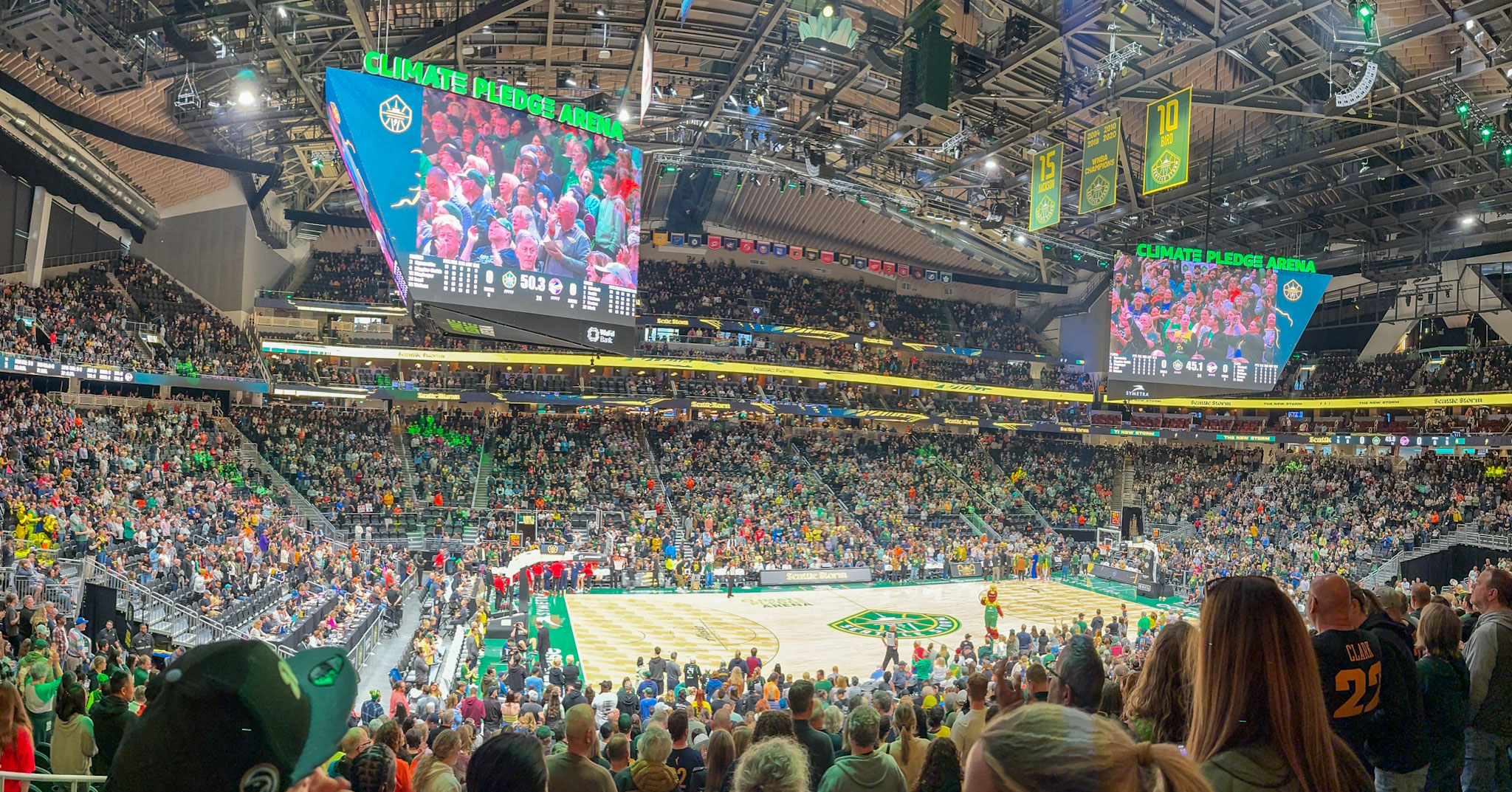 ساحة Climate Pledge مليئة بالمشجعين الذين يشاهدون مباراة Seattle Storm. تطفو شاشتان تلفزيونيتان عملاقتان فوق الملعب، تعرضان المشجعين في الساحة.