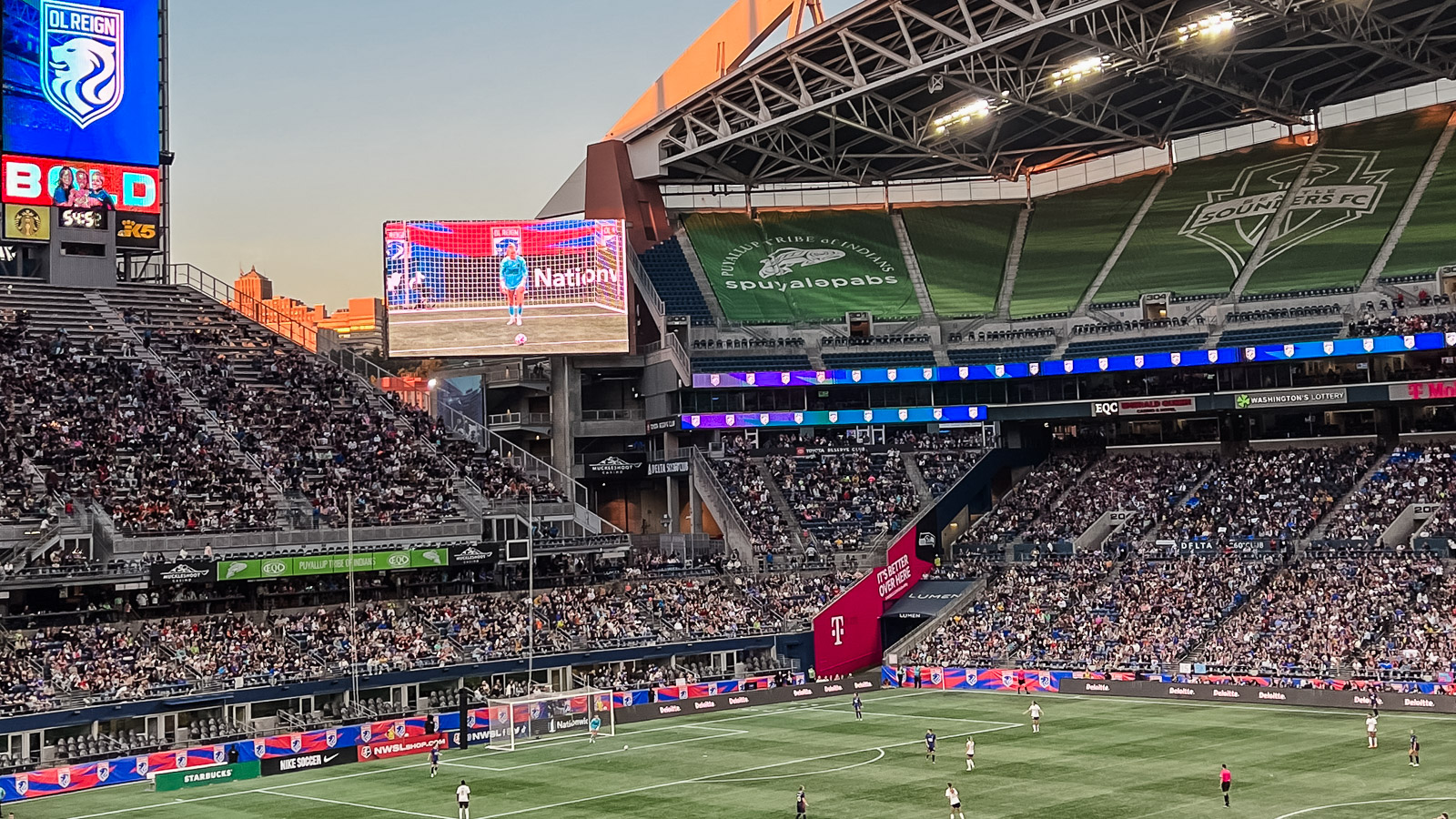 فريق Reign FC يلعب مباراة في ملعب Lumen Field. السماء تلقي ضوءًا متوهجًا على الملعب ذي العشب الأخضر. توجد شاشة تلفزيون كبيرة في منتصف يسار الصورة، تسلط الضوء على حارس المرمى في الملعب وهو على وشك تسديد ركلة.