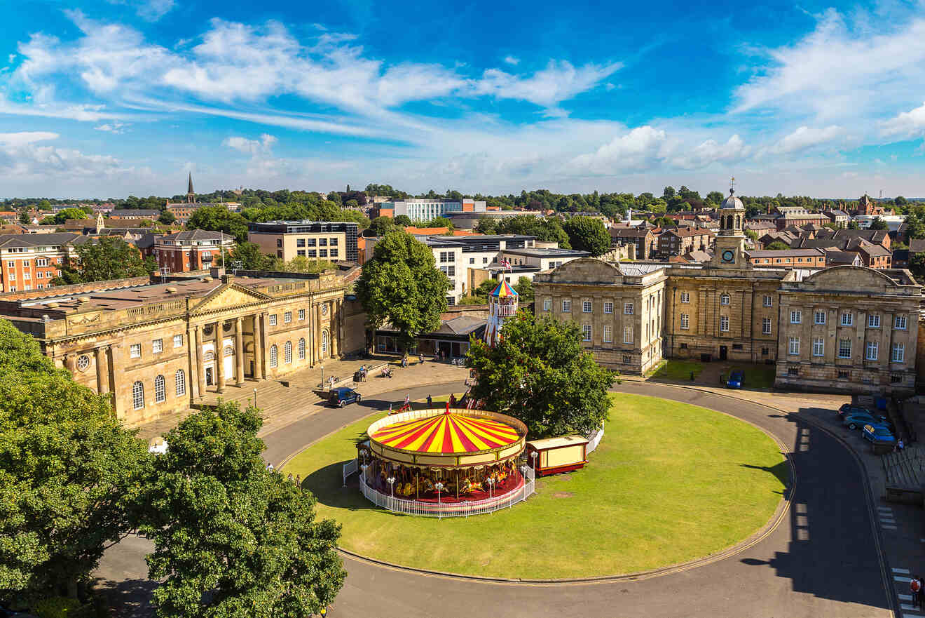 A circular building with a red and yellow striped roof sits on a grassy area surrounded by historic buildings under a blue sky with clouds.