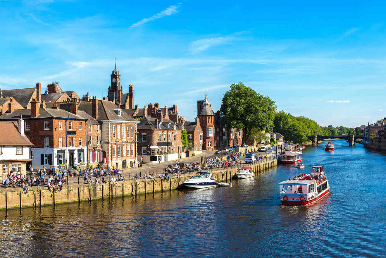 View of the River Ouse in York, England, with boats cruising along the water, people gathered along the quay, and historic brick buildings under a bright blue sky.