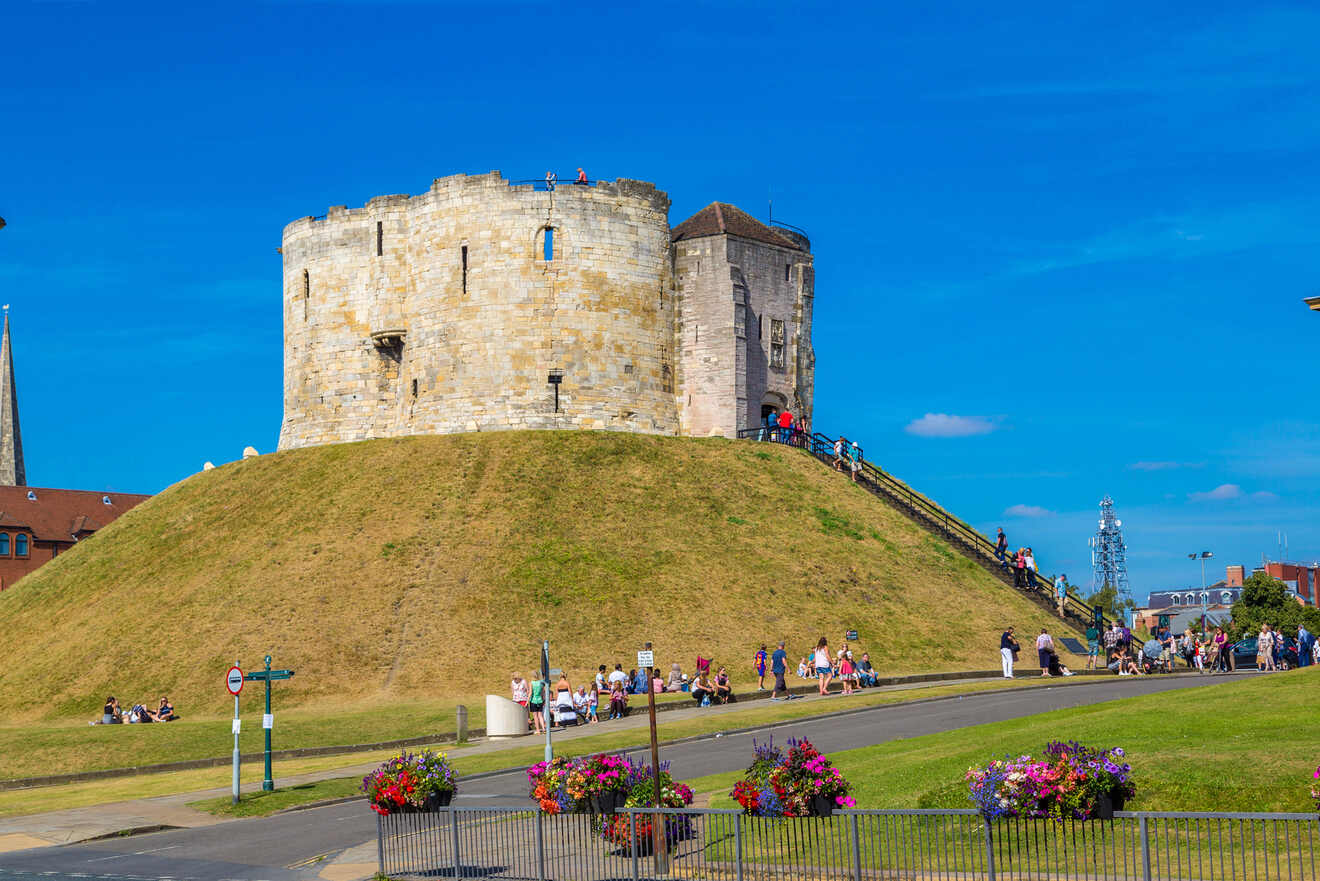 Stone tower atop a grassy mound with people walking up a path and sitting nearby; bright blue sky in the background.