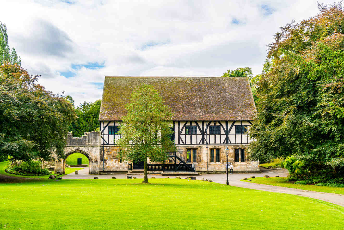 A medieval-style building with a thatched roof and timber framing is surrounded by a well-maintained lawn and trees. An arched entrance is visible to the left.