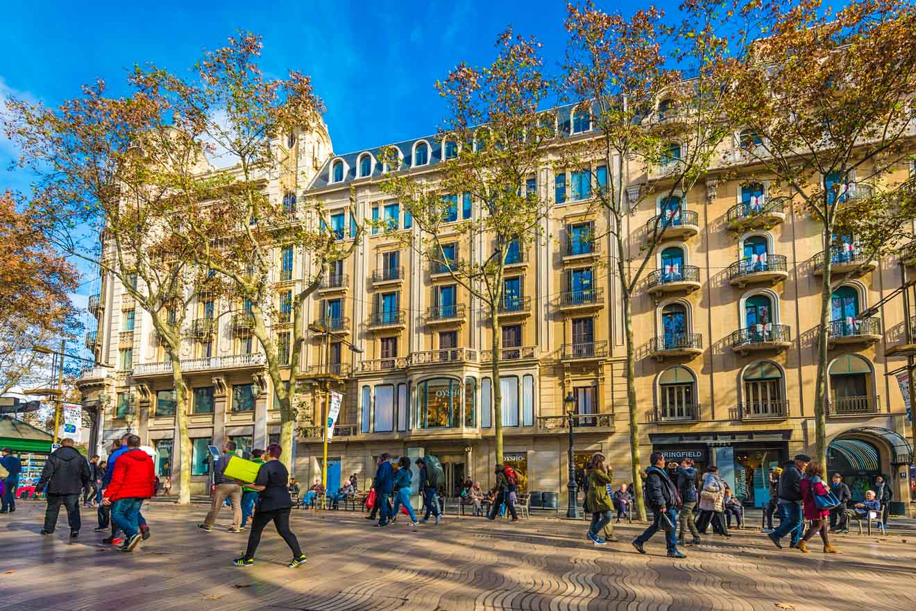 Pedestrians walking along a wide sidewalk lined with trees and historic buildings on a sunny day.