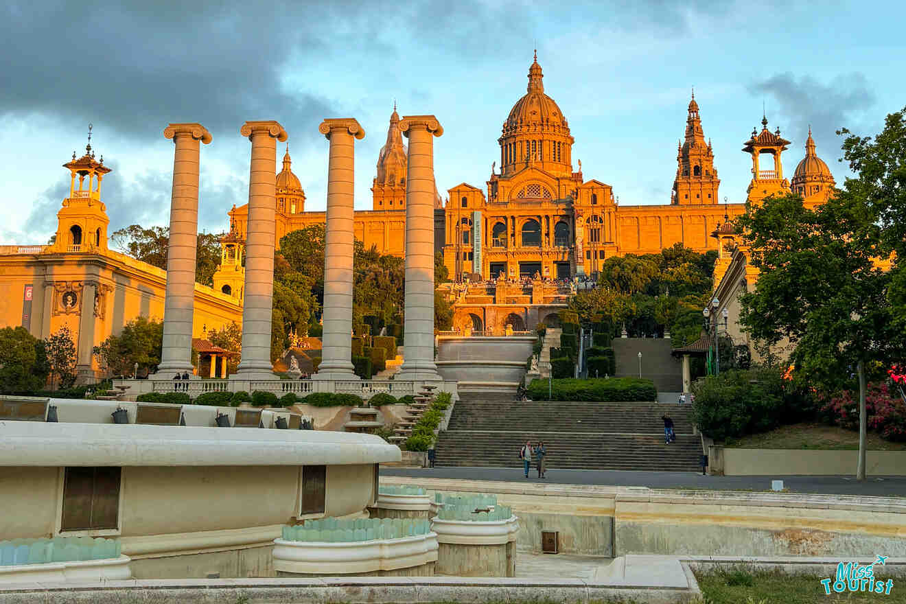 A grand palace with a dome and multiple spires sits atop a hill, illuminated by sunlight. Four tall columns and a tiered fountain are in the foreground, surrounded by steps and greenery.