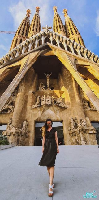 Yulia, the founder of this website, in a black dress walks in front of the Sagrada Familia's intricate facade, with sunlight illuminating the towers.