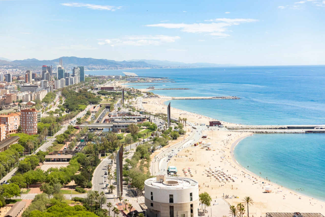 Aerial view of a coastal city showing a long beach, ocean waves, city buildings, green spaces, and mountains in the distance under a clear blue sky.