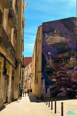 A narrow street features a large mural of a man wearing sunglasses on a building wall. Three people walk towards the mural under a clear blue sky.