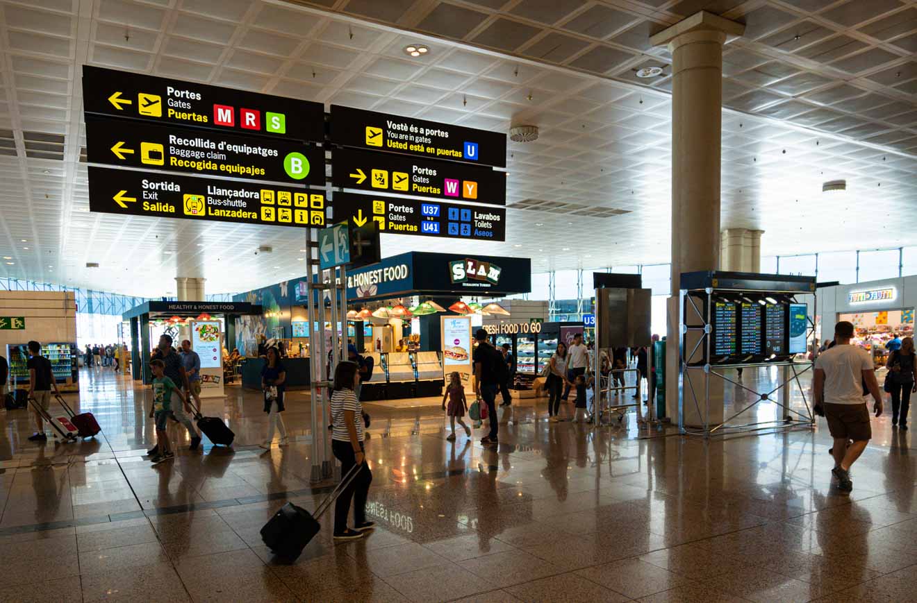 Passengers walk through a busy airport terminal with various signs overhead indicating directions to gates, baggage claim, and other services.