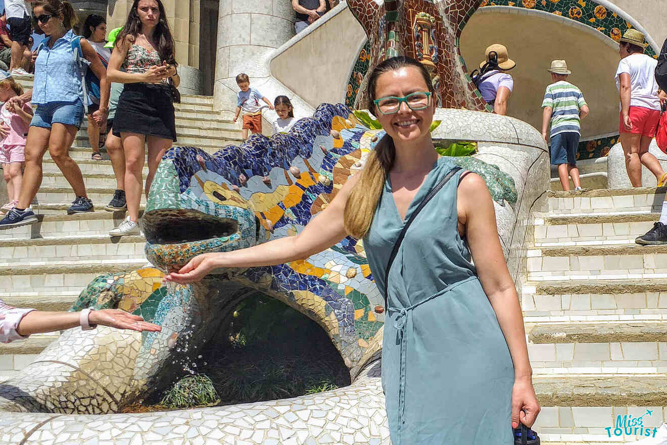 Yulia, the founder of this website, in a light blue dress poses next to a colorful mosaic lizard sculpture, with people walking down the stairs in the background.