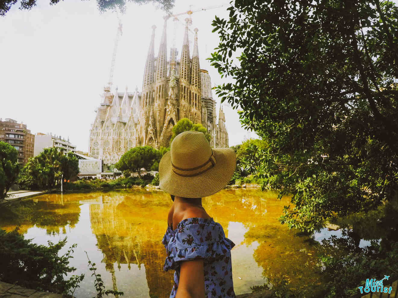 Yulia, the founder of this website, in a hat and floral dress stands by a pond, looking at the Sagrada Familia, with construction cranes visible in the background.