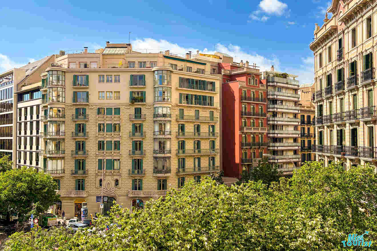 A sunny view of several multi-story residential buildings with balconies in an urban area, surrounded by leafy green trees under a blue sky with some clouds.