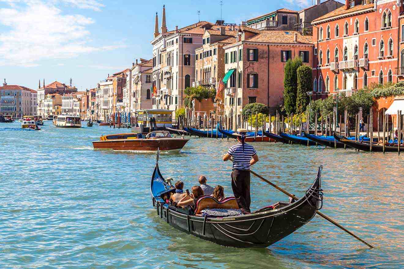 A gondolier rows a gondola with passengers along a canal in Venice, Italy, with historic buildings lining the waterway on a sunny day.