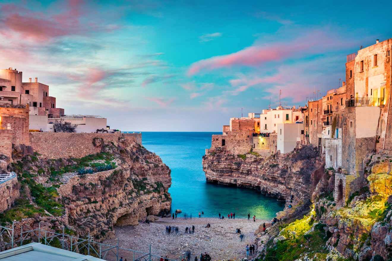 Small rocky cove with turquoise water, surrounded by cliffside buildings at sunset. People gathered on a pebble beach with the sea in the background.