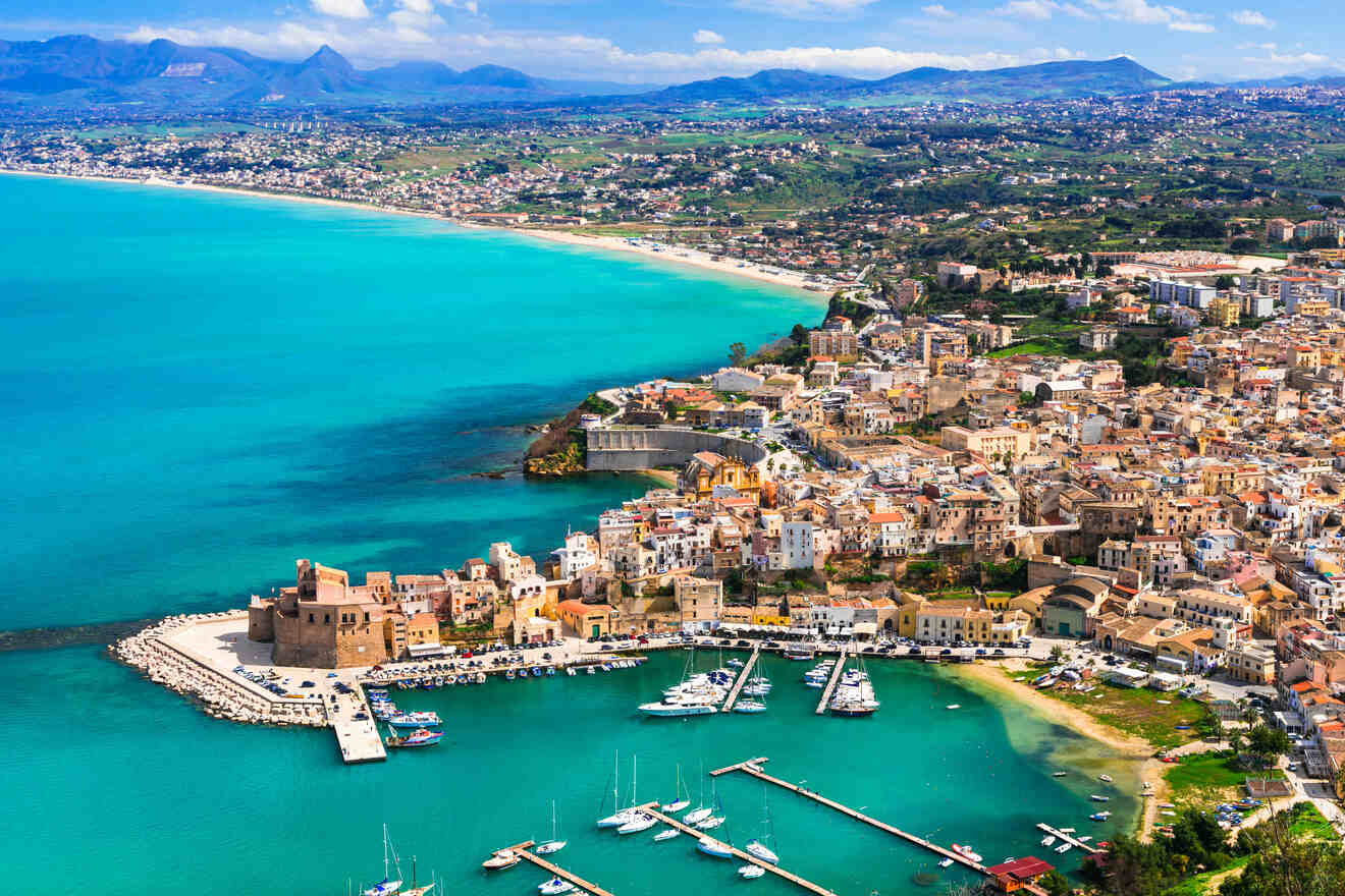 Aerial view of a coastal town with a marina, historic buildings, and a sandy beach lining a turquoise bay, set against a backdrop of green hills and distant mountains under a blue sky.