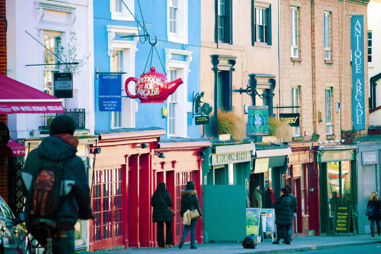 A street scene with people walking past colorful storefronts, including an antique shop and an establishment with a teapot-shaped sign hanging outside.