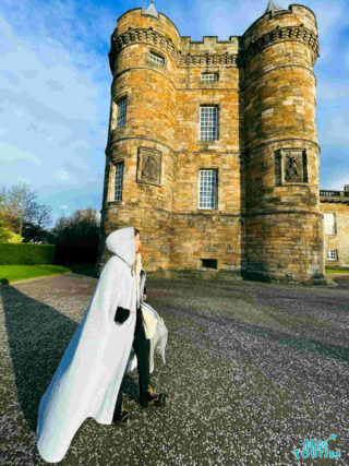 The writer of the post wearing a white hooded cape, standing in front of the historic Holyrood Palace in Edinburgh.