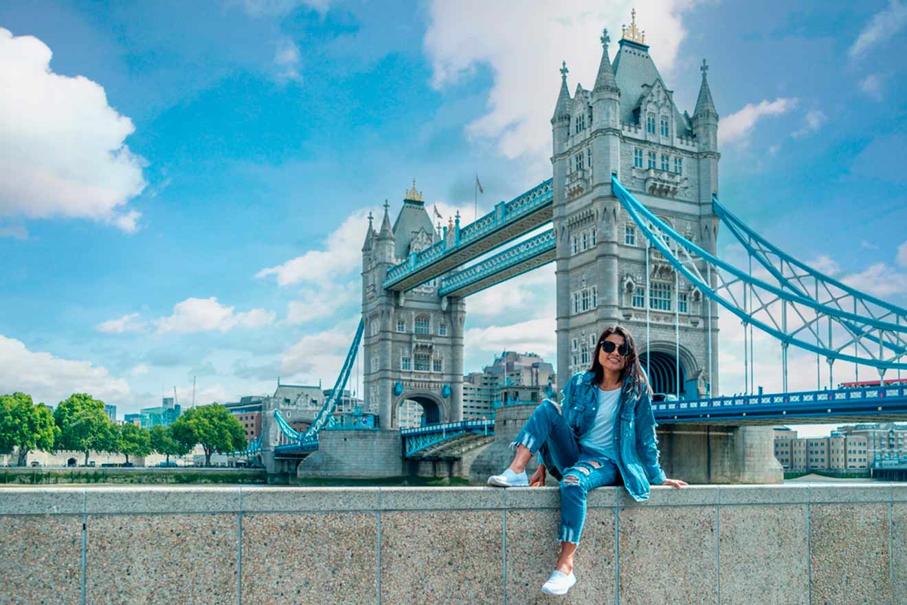 A person in casual attire sits on a ledge with the Tower Bridge in the background on a sunny day.