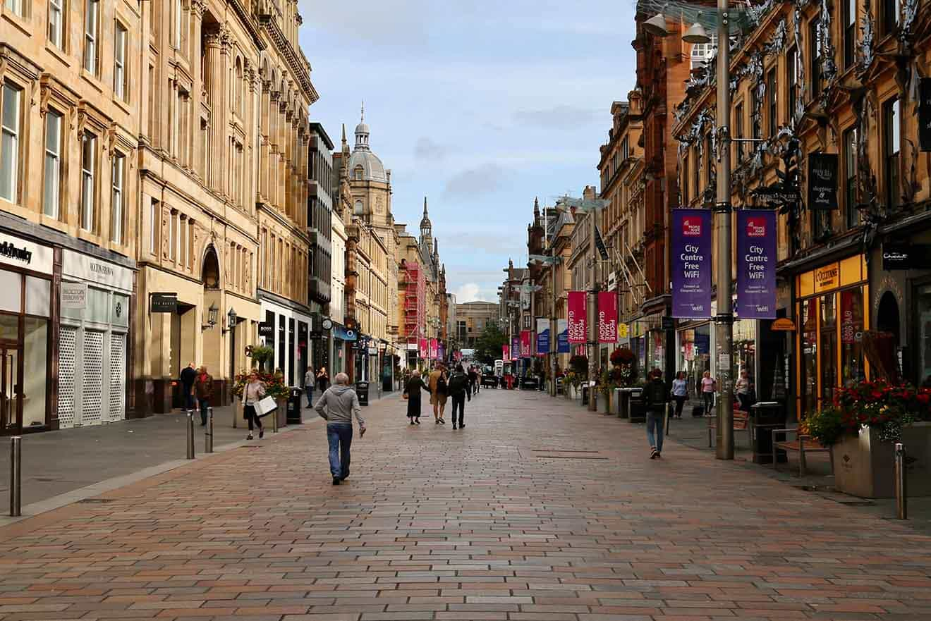 Pedestrians strolling down Buchanan Street in Glasgow, a bustling shopping thoroughfare flanked by historic architecture and modern retail outlets, under a cloudy sky.