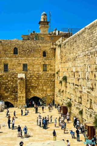 People gather at the Western Wall in Jerusalem, a significant religious site, with the ancient stone wall and the cityscape in the background under a clear blue sky.