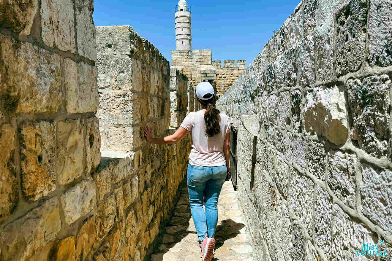 author of the post in casual attire walks along a narrow stone pathway with one hand touching the wall. A tower is visible in the background under a clear blue sky.