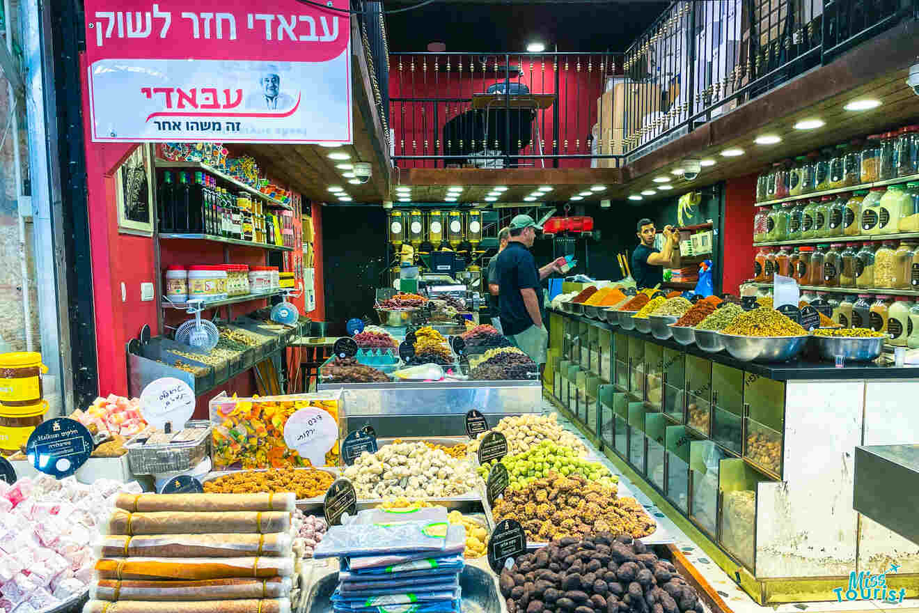 A vibrant market stall with an array of spices, dried fruits, and confections displayed in containers. Two people are seen working behind the counter. Shelves with additional jars are visible in the background.