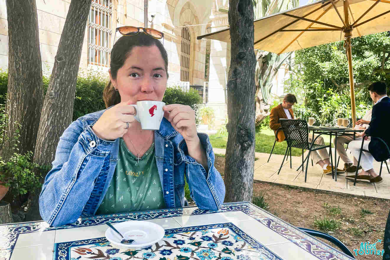 author of the posts sits at a tiled outdoor table, drinking from a white cup. Behind, two others are seated at a separate table under a canopy. Trees and greenery surround the area.