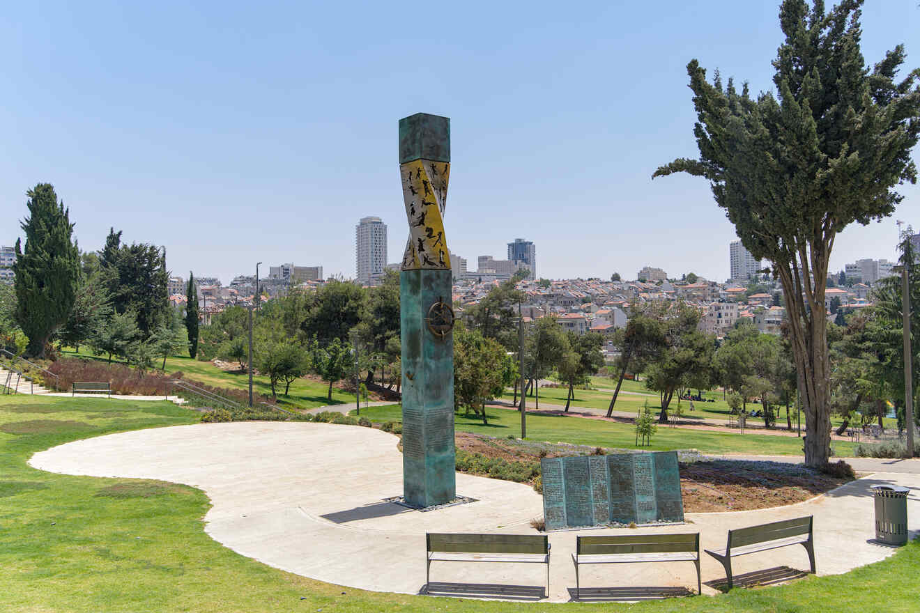 A tall outdoor sculpture stands on a paved area surrounded by grass, trees, and benches, with a cityscape in the background on a clear day.