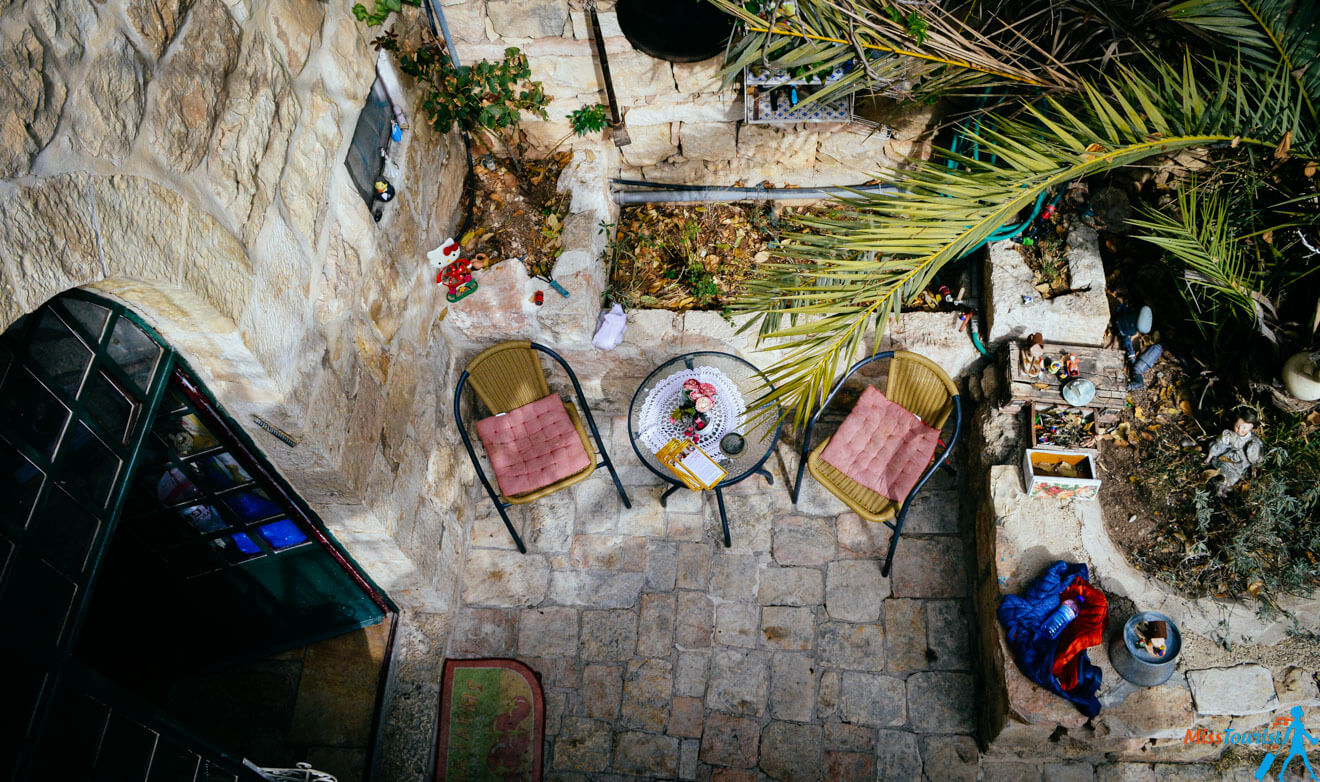 A cozy stone courtyard with two chairs, a small round table with items, surrounded by plants and a palm tree. A garden area with decorative items is visible to the right.