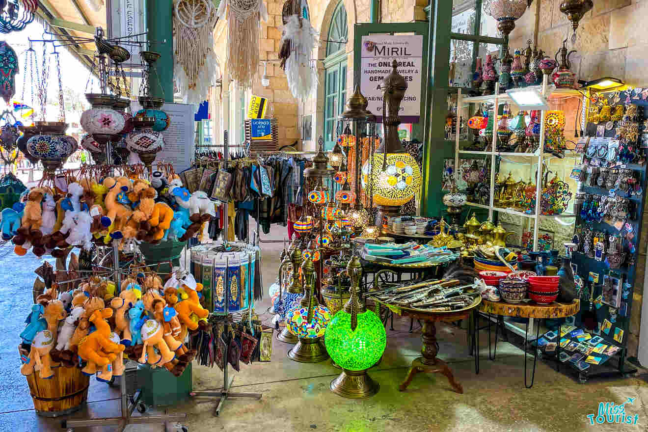 A colorful store displays various souvenirs, including stuffed camels, mosaic lamps, decorative plates, and bowls. Shelves and racks are filled with an array of items for sale.