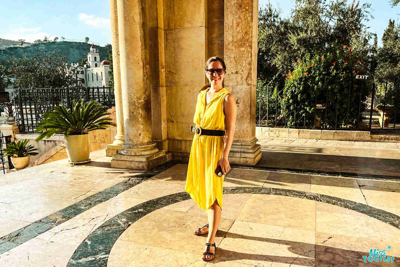 A person in a yellow dress stands in front of a large stone building with columns. There are potted plants and trees in the background. The person is smiling and holding a phone.