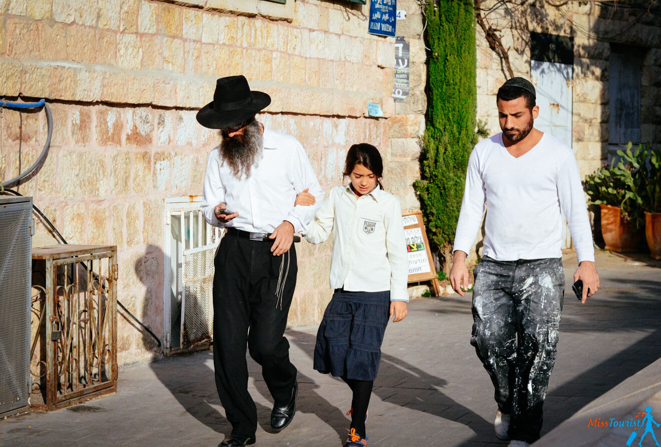 Three people walking on a sidewalk; a man in traditional dress holding a girl's arm and looking at his phone, another man in casual clothes walking beside them.