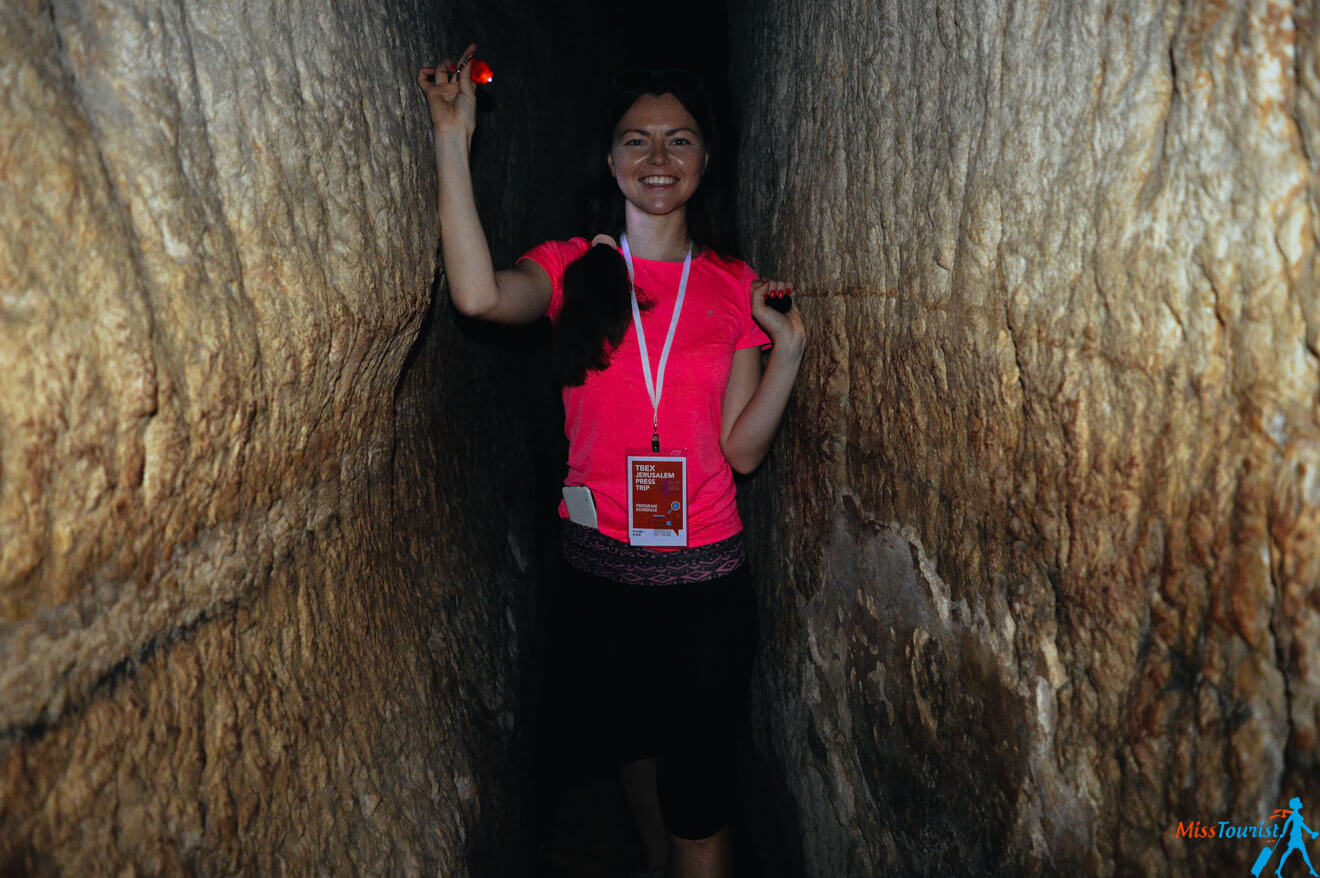 Yulia, the founder of this website, in a pink shirt and black pants stands in a narrow rock passageway, holding a small light. She wears a lanyard with a badge around her neck. The walls crowd closely beside her.