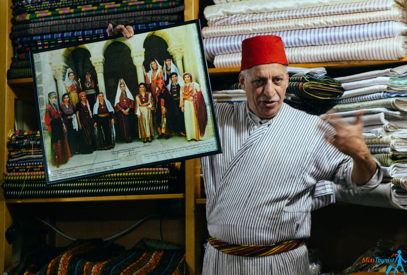 A man wearing a red fez and striped clothing holds and points to a framed photograph of a group of people in traditional attire. He stands in front of neatly stacked fabric rolls.