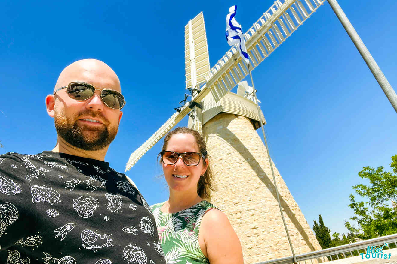 author of the posts with her husband stand in front of a stone windmill with white blades on a sunny day. The woman is wearing sunglasses and a green patterned top, while the man is wearing sunglasses and a black patterned shirt.