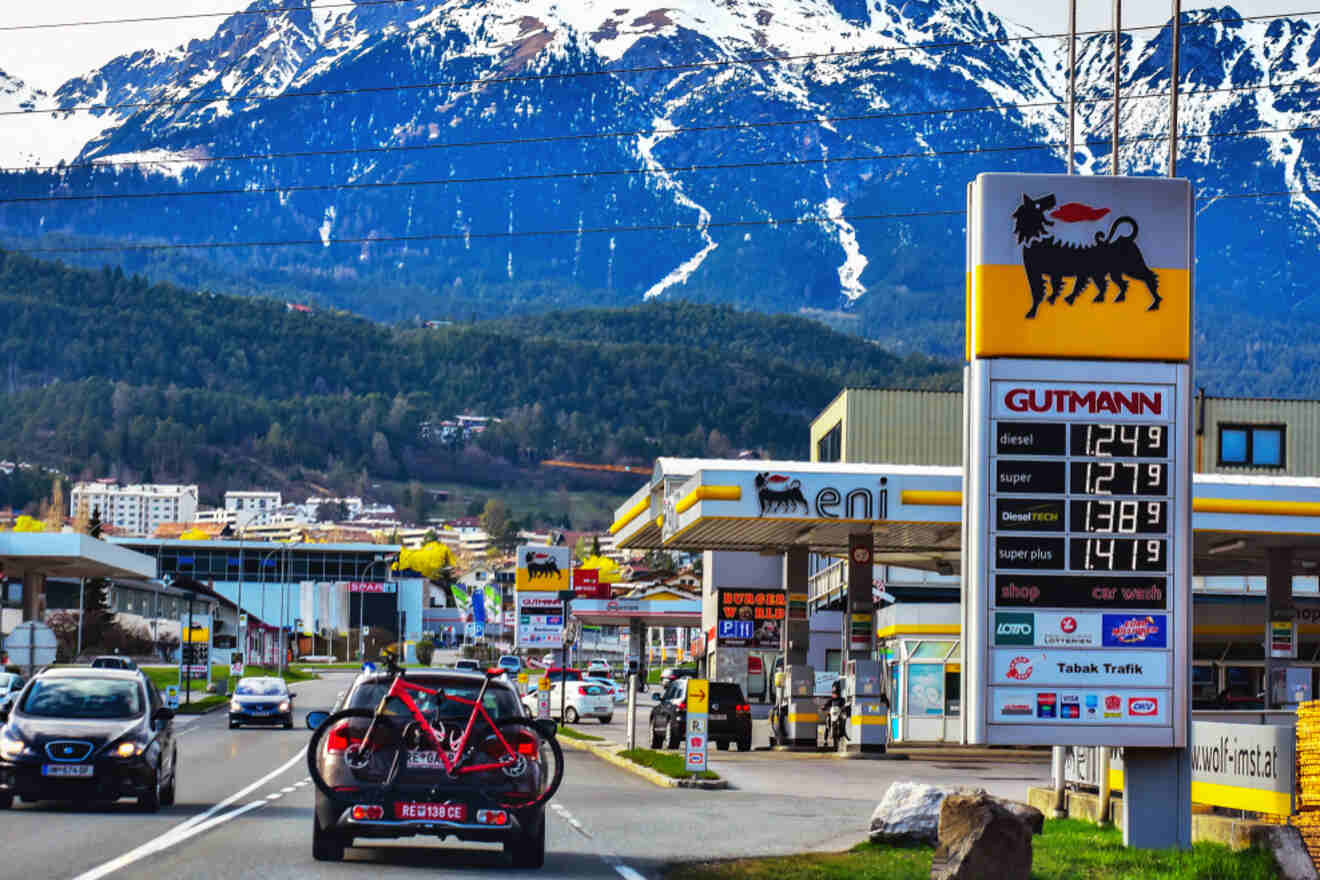 A car carrying a bicycle on its rear drives on a road near an Eni gas station. Fuel prices are displayed on a sign in the foreground, with snow-capped mountains visible in the background.