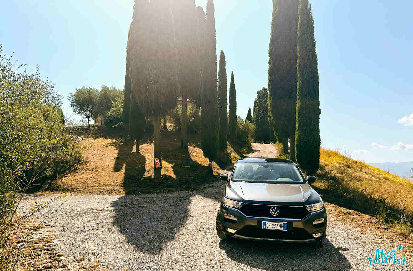 A modern car parked on a gravel road flanked by tall, dark cypress trees under a bright sun, in the picturesque Tuscan landscape.