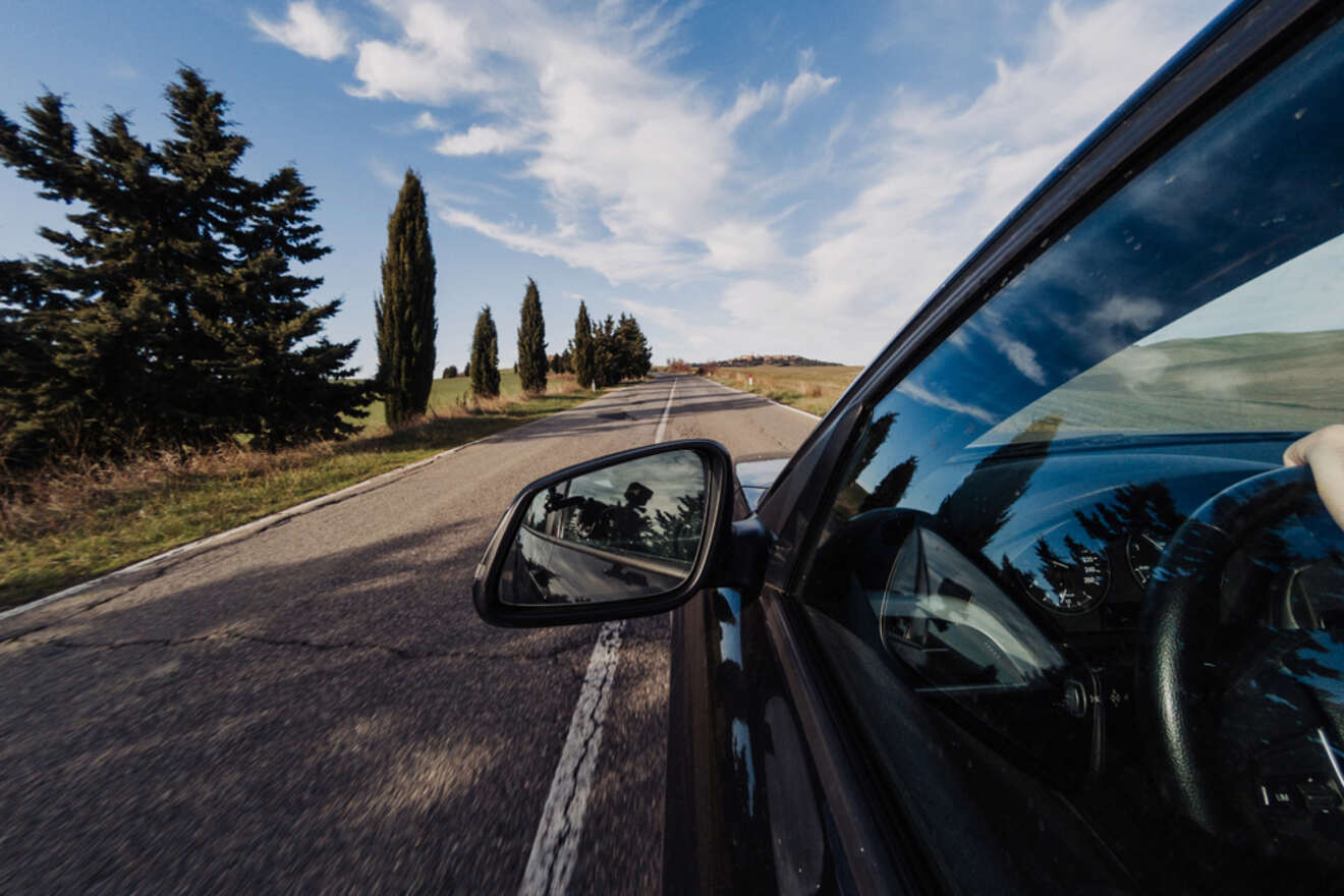 Car traveling on a straight, scenic road with trees on the side. Photo taken from the passenger side showing the side mirror and wheel in motion.