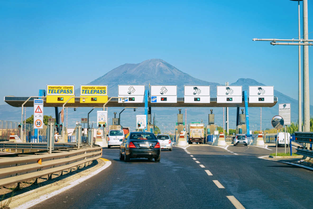 A highway toll booth with multiple lanes and signage for different payment methods, under a clear blue sky with a mountain in the background. Several vehicles are approaching or passing through.