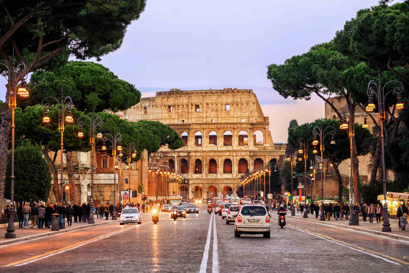 Street view of the Colosseum in Rome, Italy, with cars, motorcycles, and pedestrians under evening lights and trees lining the road.