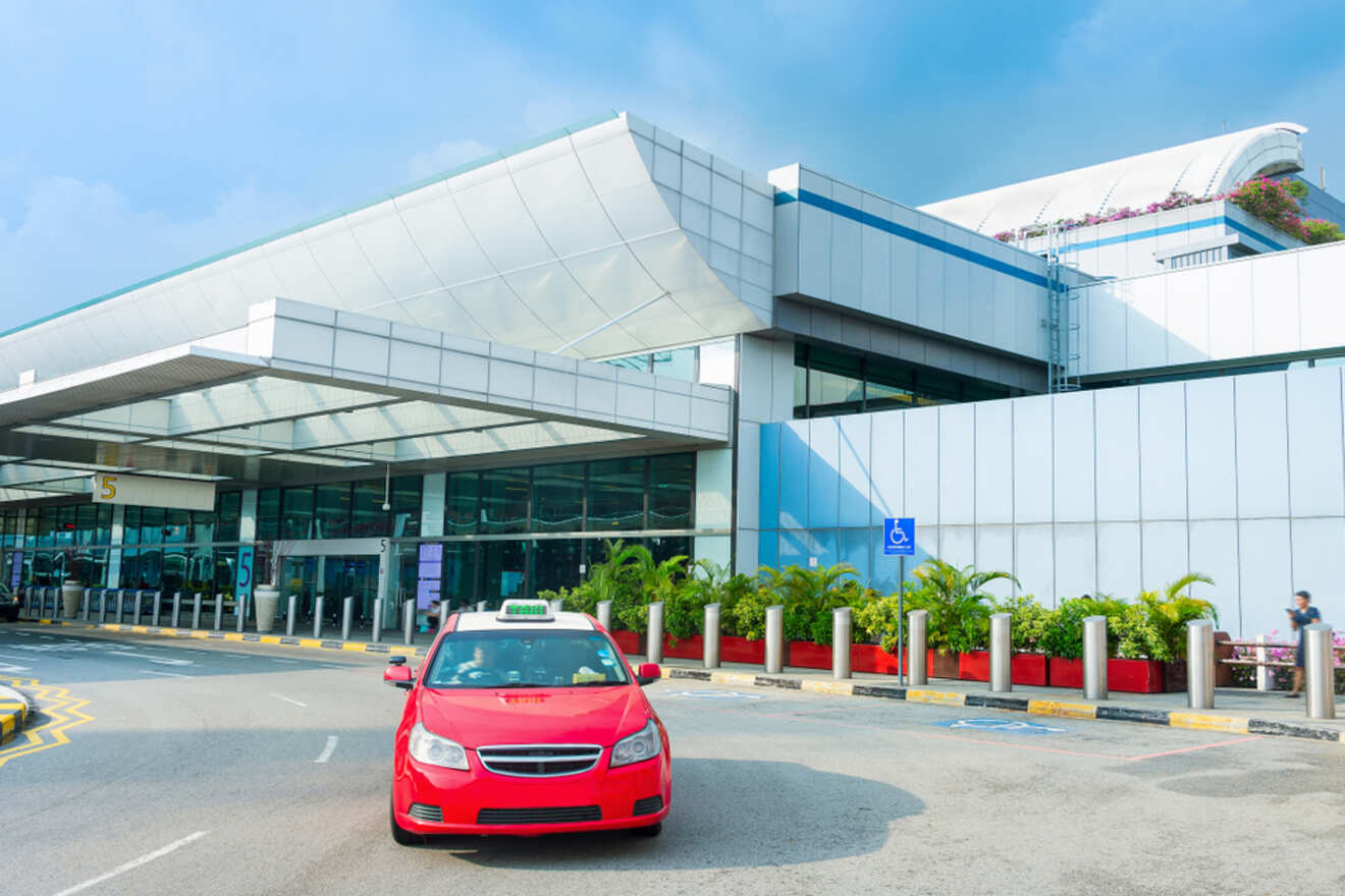 A red taxi drives past Terminal 5 of a modern airport with glass facades and white panels, under a clear blue sky.