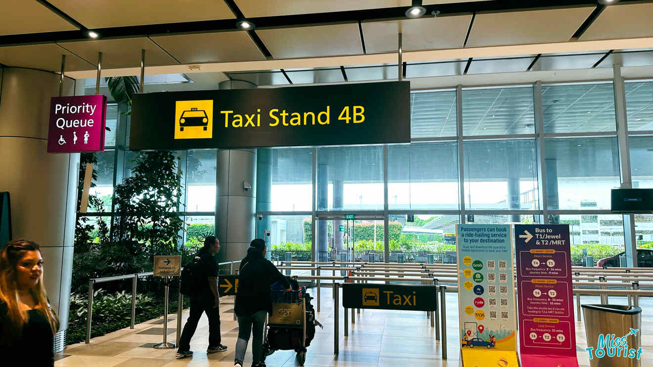 A woman with luggage walks towards the sign for Taxi Stand 4B inside an airport. There is also a sign for a Priority Queue for people with disabilities or young children.