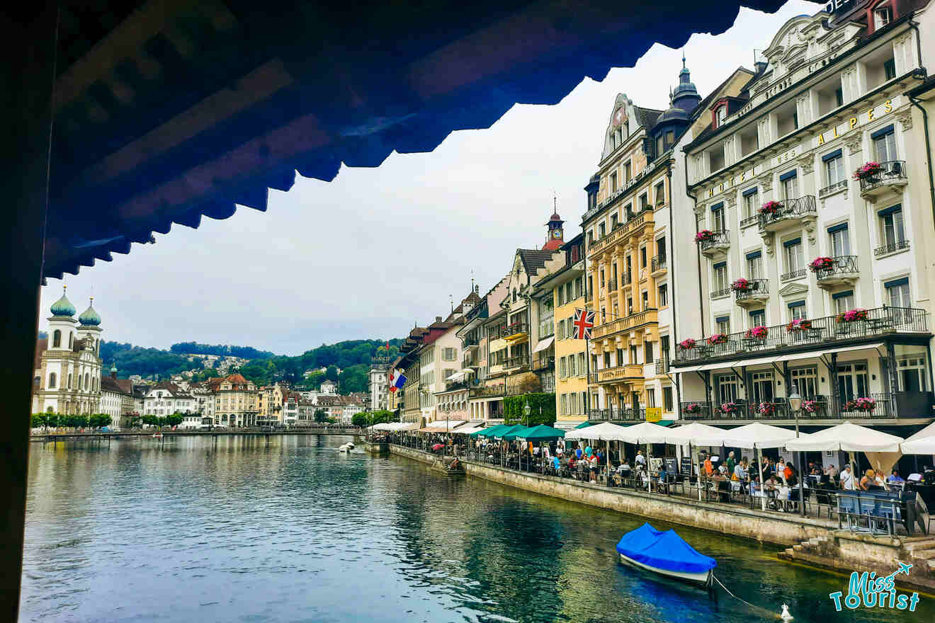 Riverside buildings with cafes and balconies line a waterfront in a European town, with hills and greenery in the background. A blue boat is moored in the water under a cloudy sky.