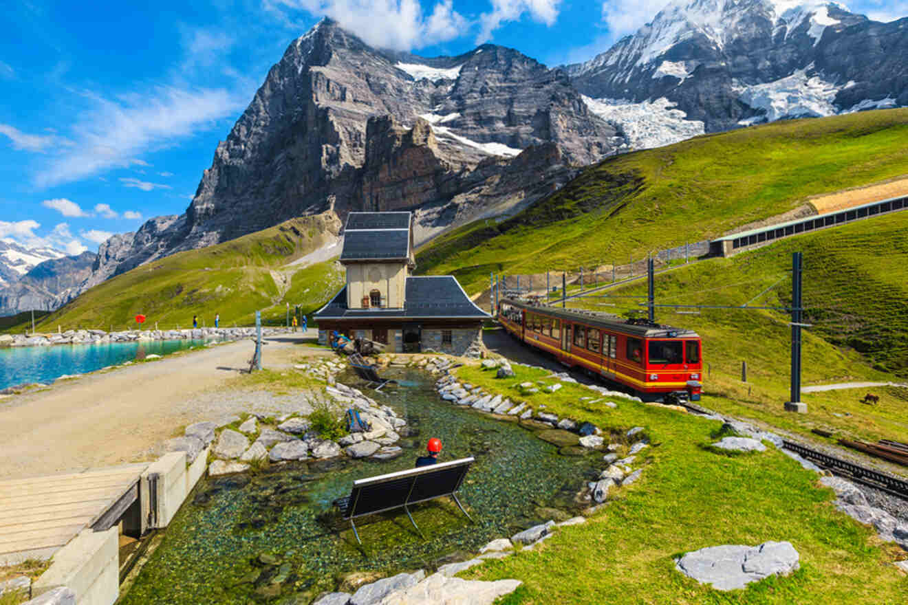 A red train passes by a small station near a clear stream with a bench, set against a backdrop of snow-capped mountains and green hills under a blue sky.